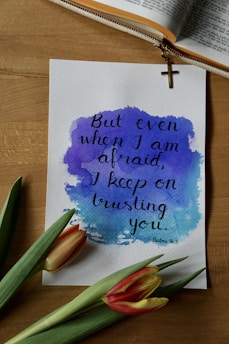 A handwritten quote on watercolor paper with a blue and purple abstract background, reading 'But even when I am afraid, I keep on trusting you. Psalm 56:3'. The paper is placed on a wooden surface near a pair of tulips and an open book with a zippered cover featuring a hanging cross.