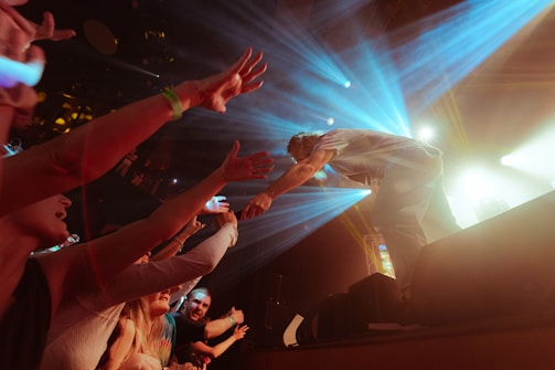 A performer on stage reaches out to a crowd of enthusiastic fans, who extend their arms towards the stage. Bright stage lights create a dynamic atmosphere with beams of blue and yellow light.