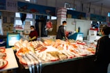 A fish market stall with a variety of fish and seafood displayed on a large table covered with ice. There are three people: one person standing behind the stall, another person possibly a customer, and another person arranging the goods. The setting appears to be indoors with bright lighting, and the interior has signs and posters related to fish hanging on the walls. The individuals are wearing face masks.
