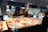 A fish market stall with a variety of fish and seafood displayed on a large table covered with ice. There are three people: one person standing behind the stall, another person possibly a customer, and another person arranging the goods. The setting appears to be indoors with bright lighting, and the interior has signs and posters related to fish hanging on the walls. The individuals are wearing face masks.