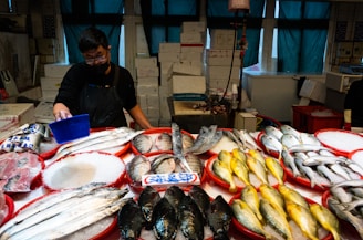 A fish market stall with various types of fresh fish arranged on red trays filled with ice. A person wearing a dark apron and mask is tending to the stall, using a blue scoop. There are numerous boxes stacked behind the counter. The lighting is dim, contributing to a slightly moody ambiance, with green curtains partially covering the windows in the background.