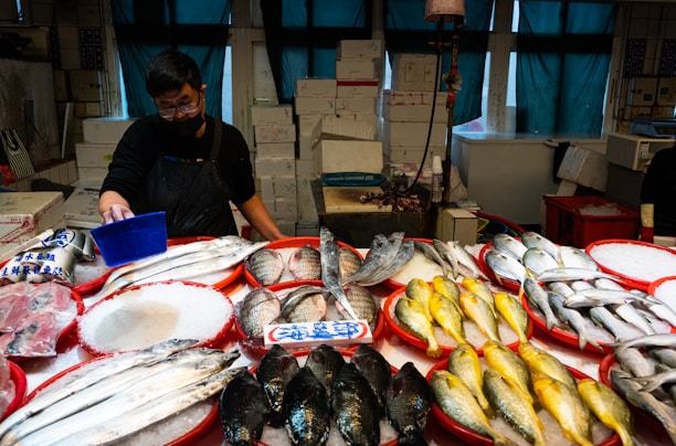 A fish market stall with various types of fresh fish arranged on red trays filled with ice. A person wearing a dark apron and mask is tending to the stall, using a blue scoop. There are numerous boxes stacked behind the counter. The lighting is dim, contributing to a slightly moody ambiance, with green curtains partially covering the windows in the background.
