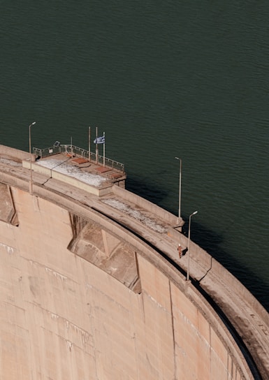 A large concrete dam curves dramatically, set against a backdrop of green water. On top of the dam, there is a small platform with a flagpole and a flag. A couple of street lamps line the walkway along the dam.