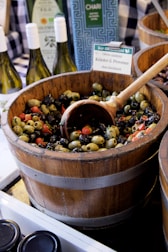 a wooden bucket filled with olives next to bottles of wine
