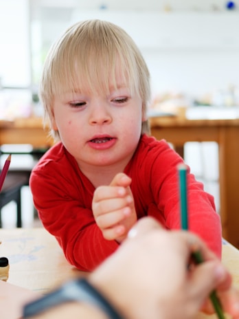 A child with blond hair wearing a red shirt is engaged in an activity at a table. One of their hands is reaching towards a pencil being held by another person. The setting is a bright room with a wooden table and blurred objects in the background.