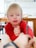 A child with blond hair wearing a red shirt is engaged in an activity at a table. One of their hands is reaching towards a pencil being held by another person. The setting is a bright room with a wooden table and blurred objects in the background.