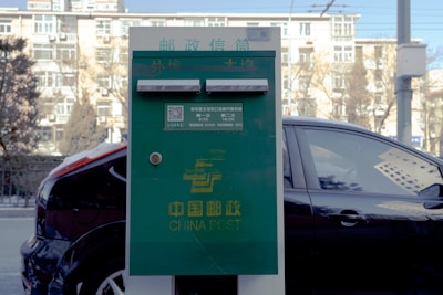 A green China Post mailbox stands on the sidewalk with its distinct yellow logo and text. In the background, a black car is parked and an apartment building with white balconies and windows is visible. The setting seems urban, and the image is taken during the day.