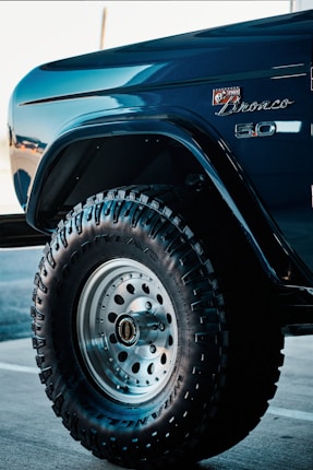 A close-up view of a vehicle's front fender and wheel featuring off-road tires with prominent treads. The car is a Bronco model, indicated by the badge on the body, finished in a glossy, deep blue color. The tire displays the Goodyear brand along with Wrangler lettering, set against a metallic wheel hub.