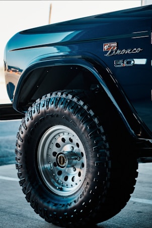 A close-up view of a vehicle's front fender and wheel featuring off-road tires with prominent treads. The car is a Bronco model, indicated by the badge on the body, finished in a glossy, deep blue color. The tire displays the Goodyear brand along with Wrangler lettering, set against a metallic wheel hub.