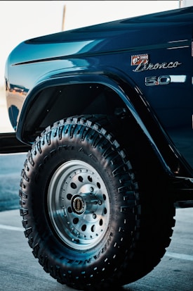 A close-up view of a vehicle's front fender and wheel featuring off-road tires with prominent treads. The car is a Bronco model, indicated by the badge on the body, finished in a glossy, deep blue color. The tire displays the Goodyear brand along with Wrangler lettering, set against a metallic wheel hub.