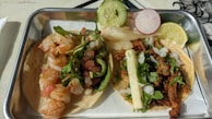 A meal featuring two types of tacos in a rectangular metal tray. One taco contains sautéed shrimp topped with greens and diced tomatoes, garnished with a slice of cucumber and a radish slice. The second taco includes seasoned meat with diced onions, cilantro, and a strip of grilled onion, garnished with a lime wedge.