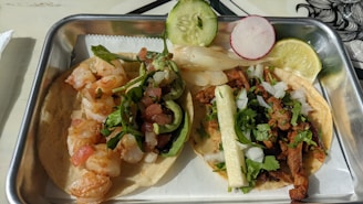 A meal featuring two types of tacos in a rectangular metal tray. One taco contains sautéed shrimp topped with greens and diced tomatoes, garnished with a slice of cucumber and a radish slice. The second taco includes seasoned meat with diced onions, cilantro, and a strip of grilled onion, garnished with a lime wedge.