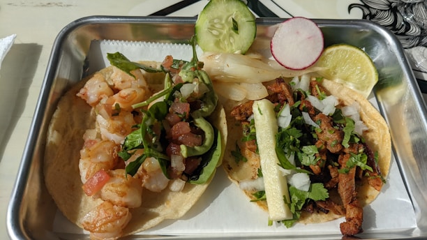 A meal featuring two types of tacos in a rectangular metal tray. One taco contains sautéed shrimp topped with greens and diced tomatoes, garnished with a slice of cucumber and a radish slice. The second taco includes seasoned meat with diced onions, cilantro, and a strip of grilled onion, garnished with a lime wedge.