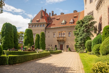 A historic castle with an ornate facade and red-tiled roof is surrounded by a well-maintained garden. Tall, manicured shrubs and lush greenery line the cobblestone pathway leading up to the entrance. Visitors are seen relaxing on benches and enjoying the serene atmosphere.