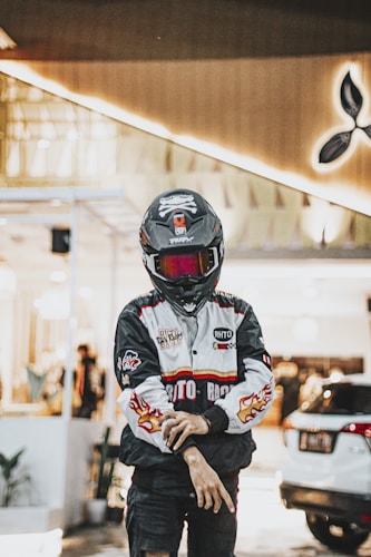 A person wearing a black motorcycle helmet and a branded jacket featuring logos and flame designs stands in front of a background that includes a building with bright lights and a partially visible car.