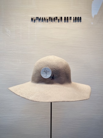A beige wide-brimmed hat is displayed on a stand against a neutral background. Above the hat, the text 'HUTMANUFAKTUR SEIT 1903' is visible, suggesting a hat manufacturer established in 1903.
