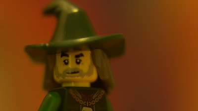 A Lego figure resembling a wizard with a pointy green hat and gray beard is depicted against a blurred warm-toned background.
