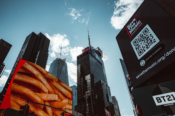 Skyscrapers rise against a partly cloudy sky, with large digital billboards displaying advertisements including an image of fries and a Save the Music Foundation QR code.