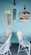 A light blue wall features an eclectic arrangement of decor items including a mix of painted and rustic wooden panels, geometric shapes, and a sign that reads 'Do What You Love.' There are two white wicker chairs placed against the wall, and above them are shelves filled with books and small items. A single sconce light fixture is mounted on the wall.