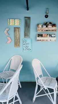 A light blue wall features an eclectic arrangement of decor items including a mix of painted and rustic wooden panels, geometric shapes, and a sign that reads 'Do What You Love.' There are two white wicker chairs placed against the wall, and above them are shelves filled with books and small items. A single sconce light fixture is mounted on the wall.