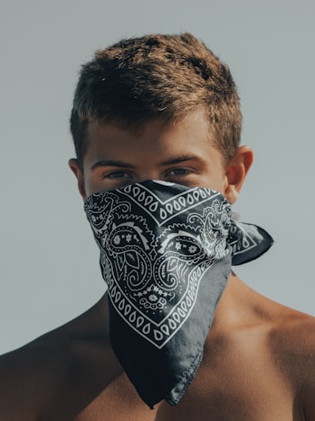 A person stands with a neutral background, wearing a patterned bandana that covers the lower half of the face. The person has short, brown hair and is shirtless.