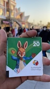 A hand is holding a card with a value of 20 AED. The card features a cartoon rabbit wearing a blue jacket and green scarf. The background shows a busy outdoor setting with numerous people and an illuminated building.
