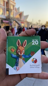 A hand is holding a card with a value of 20 AED. The card features a cartoon rabbit wearing a blue jacket and green scarf. The background shows a busy outdoor setting with numerous people and an illuminated building.