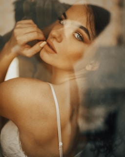 A woman with a contemplative expression looks over her shoulder. Her skin is softly lit, and she wears a thin strap white garment. Her eyes are accentuated with makeup, and her long dark hair falls loosely. The background is blurred, lending a dreamy quality to the scene.