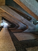 A corridor with a geometric architectural design featuring large concrete beams creating an angular pattern along the ceiling and wall. The floor is covered with a mosaic of small square tiles in a combination of beige and dark colors.