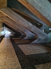 A corridor with a geometric architectural design featuring large concrete beams creating an angular pattern along the ceiling and wall. The floor is covered with a mosaic of small square tiles in a combination of beige and dark colors.