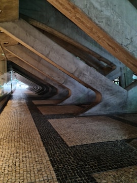 A corridor with a geometric architectural design featuring large concrete beams creating an angular pattern along the ceiling and wall. The floor is covered with a mosaic of small square tiles in a combination of beige and dark colors.
