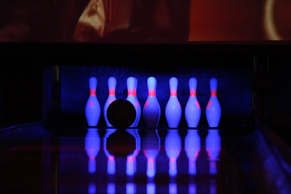 A set of bowling pins and a bowling ball are illuminated by neon blue and pink lights in a dimly lit bowling alley. The pins are positioned at the end of a lane, casting reflections on the glossy surface.