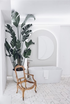 A modern bathroom with a large white bathtub, an ornate vintage chair, and a tall potted plant with large green leaves. The room features a geometric wall decoration above the bathtub and a soft, indirect lighting setup.
