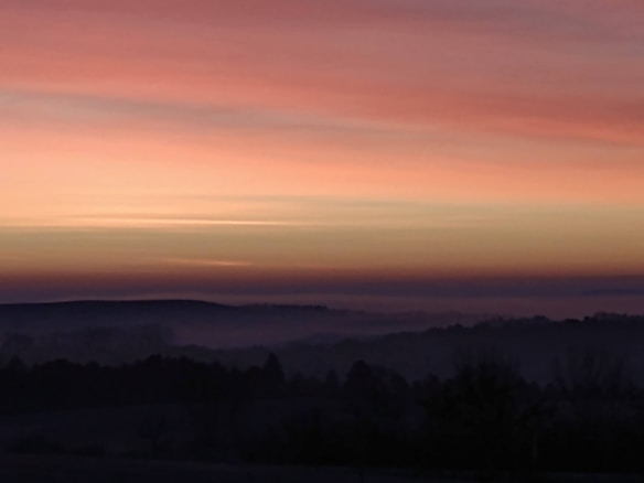 A serene landscape featuring a soft gradient sky transitioning from warm shades of pink and orange to the darker horizon. The silhouettes of hills and trees are gently enveloped in mist, adding depth and mystery.
