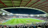A large football stadium filled with spectators. The field is brightly lit, and two teams are positioned on the pitch. The stadium is covered, with a visible roof structure and stands filled with fans. Scoreboards are visible displaying game information.