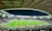 A large football stadium filled with spectators. The field is brightly lit, and two teams are positioned on the pitch. The stadium is covered, with a visible roof structure and stands filled with fans. Scoreboards are visible displaying game information.