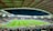 A large football stadium filled with spectators. The field is brightly lit, and two teams are positioned on the pitch. The stadium is covered, with a visible roof structure and stands filled with fans. Scoreboards are visible displaying game information.