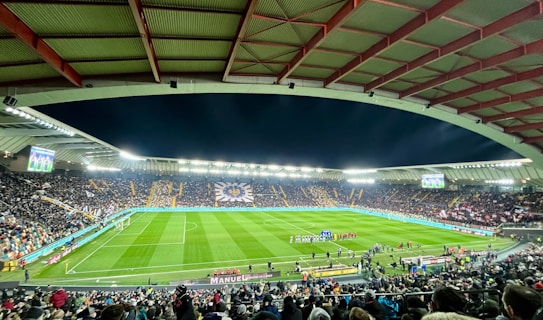 A large football stadium filled with spectators. The field is brightly lit, and two teams are positioned on the pitch. The stadium is covered, with a visible roof structure and stands filled with fans. Scoreboards are visible displaying game information.
