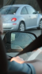 A view from inside a car showing a side mirror reflecting another vehicle. The reflection includes an image of a person driving and the text 'objects in mirror are closer than they appear.' A silver car with red taillights drives alongside.