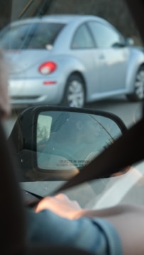 A view from inside a car showing a side mirror reflecting another vehicle. The reflection includes an image of a person driving and the text 'objects in mirror are closer than they appear.' A silver car with red taillights drives alongside.