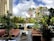 A lush outdoor seating area with modern furniture, surrounded by a variety of green plants and palm trees. The scene captures a mix of contemporary style with natural elements, set against a backdrop of tall buildings and a partly cloudy sky.