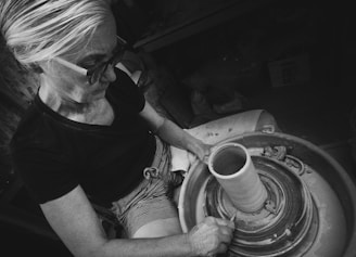 An individual is focused on shaping clay on a pottery wheel. The environment suggests a workshop setting. The person is wearing glasses and is seated, using their hands skillfully on the rotating clay. The monochromatic tone adds an artistic and classic feel.