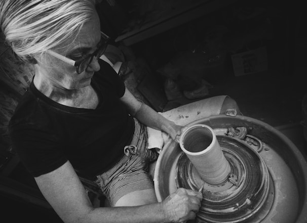 An individual is focused on shaping clay on a pottery wheel. The environment suggests a workshop setting. The person is wearing glasses and is seated, using their hands skillfully on the rotating clay. The monochromatic tone adds an artistic and classic feel.