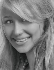 A close-up black and white photograph of a young woman with a friendly smile. Her blonde hair falls naturally around her face, and her eyes are looking slightly to the side, creating a cheerful expression.