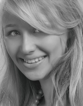 A close-up black and white photograph of a young woman with a friendly smile. Her blonde hair falls naturally around her face, and her eyes are looking slightly to the side, creating a cheerful expression.