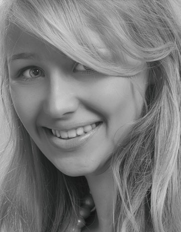 A close-up black and white photograph of a young woman with a friendly smile. Her blonde hair falls naturally around her face, and her eyes are looking slightly to the side, creating a cheerful expression.