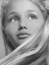 A black and white portrait of a young person with long, flowing hair. The expression is thoughtful, with eyes gazing upward. Fine details are visible in the hair and facial features, conveying a sense of softness.