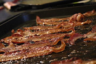 Crispy bacon strips lie on a greasy, dark-colored cooking surface. The bacon has a golden-brown hue with visible fat sizzling.