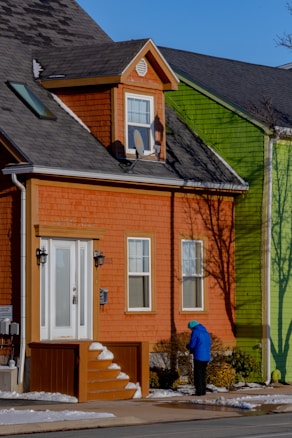 A two-story house with orange siding, white trim, and a satellite dish is positioned next to a vibrant green building. A person wearing a bright blue jacket and teal beanie stands near the entrance, surrounded by a small amount of snow on the ground and steps.