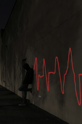 A person wearing a hoodie leans against a dimly lit wall at night. A bright red neon-style graphic resembling an electrocardiogram line is displayed on the wall beside them, casting a stark contrast against the dark surroundings.